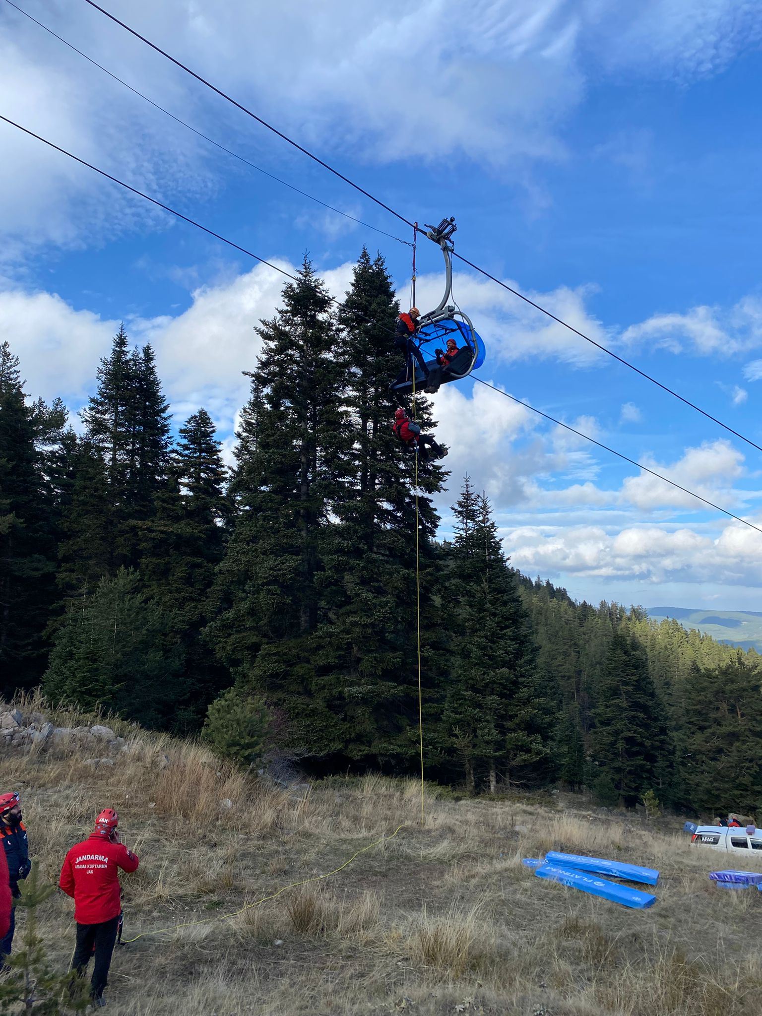A passenger being lowered by rope from a chairlift chair to the ground, with a JAK team below