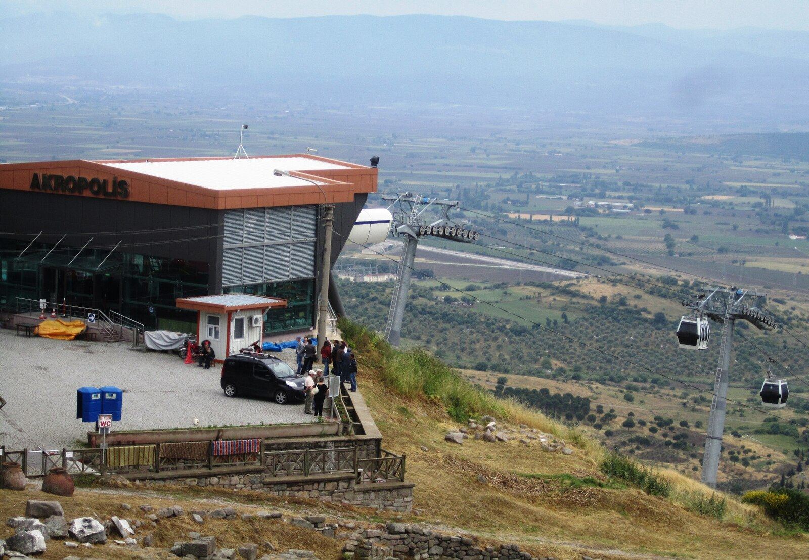Tepede Akropolis yazılı teleferik zirve istasyonu, hattın direkleri ve ovaya inen iki kabin