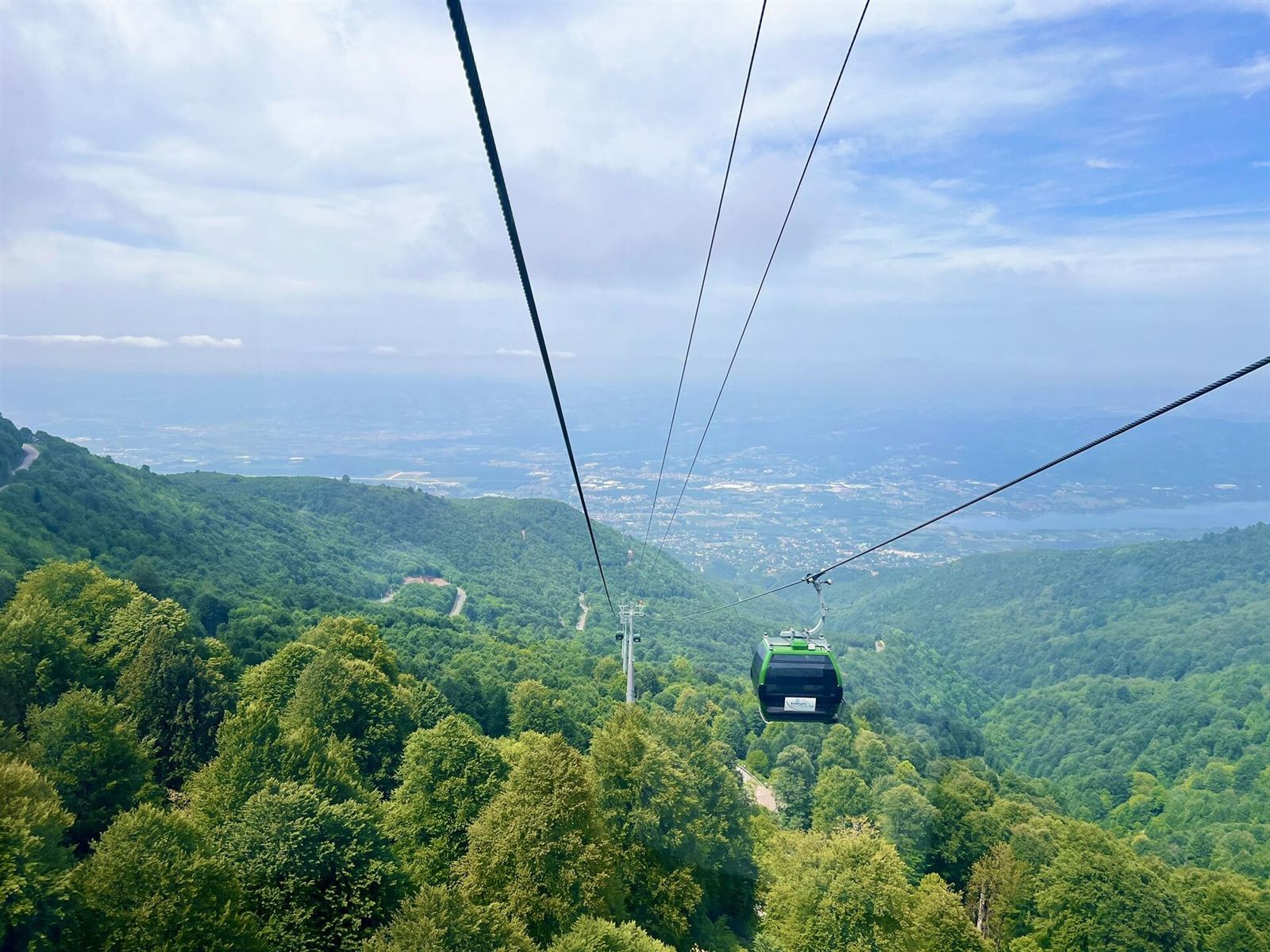 Kartepe gondol hattının yeşil kabini ormanın üzerinde; arka planda Sapanca Gölü ve ova
